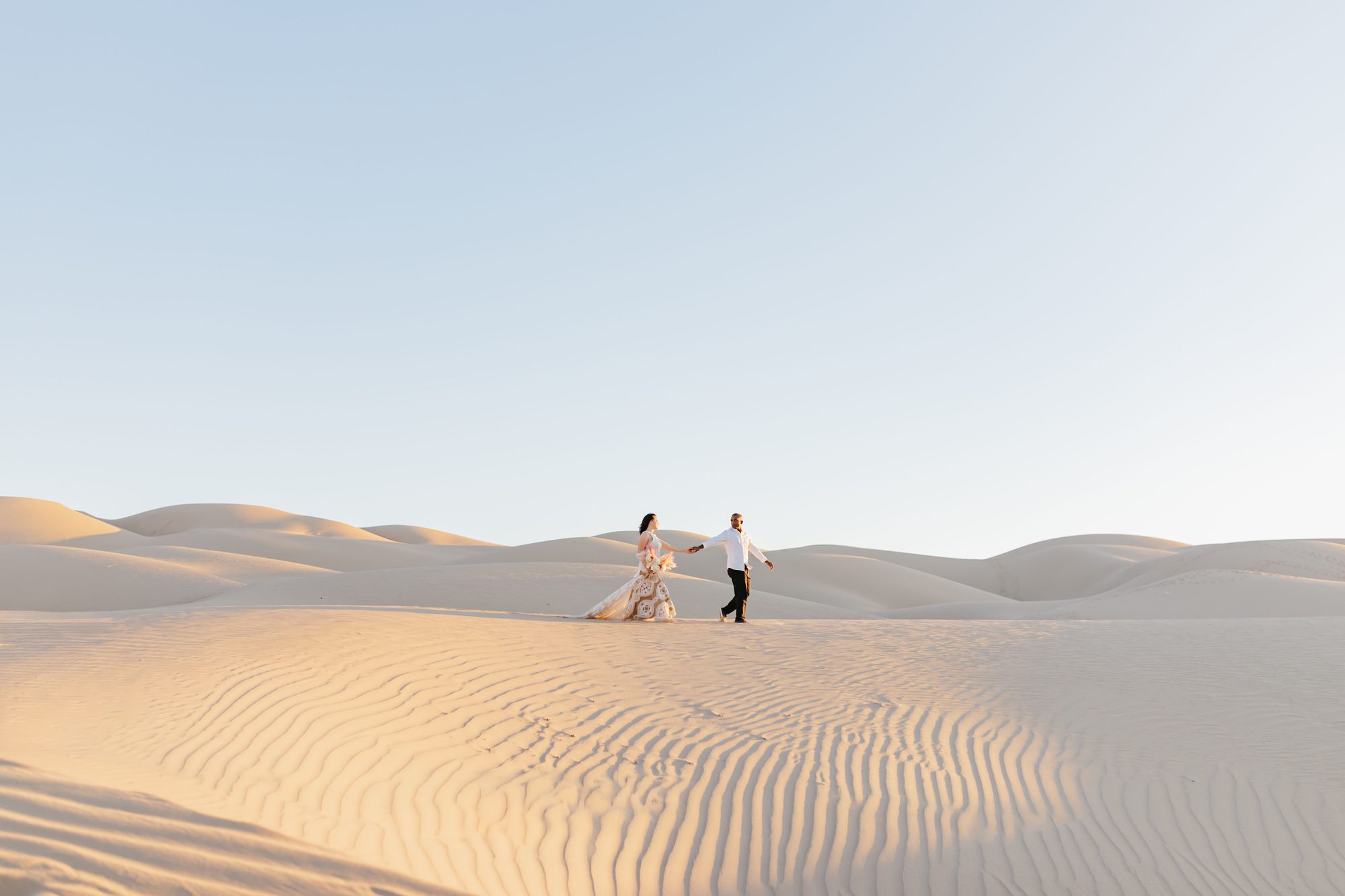 Couple walking hand in hand across golden sand dunes at sunset during destination wedding portraits