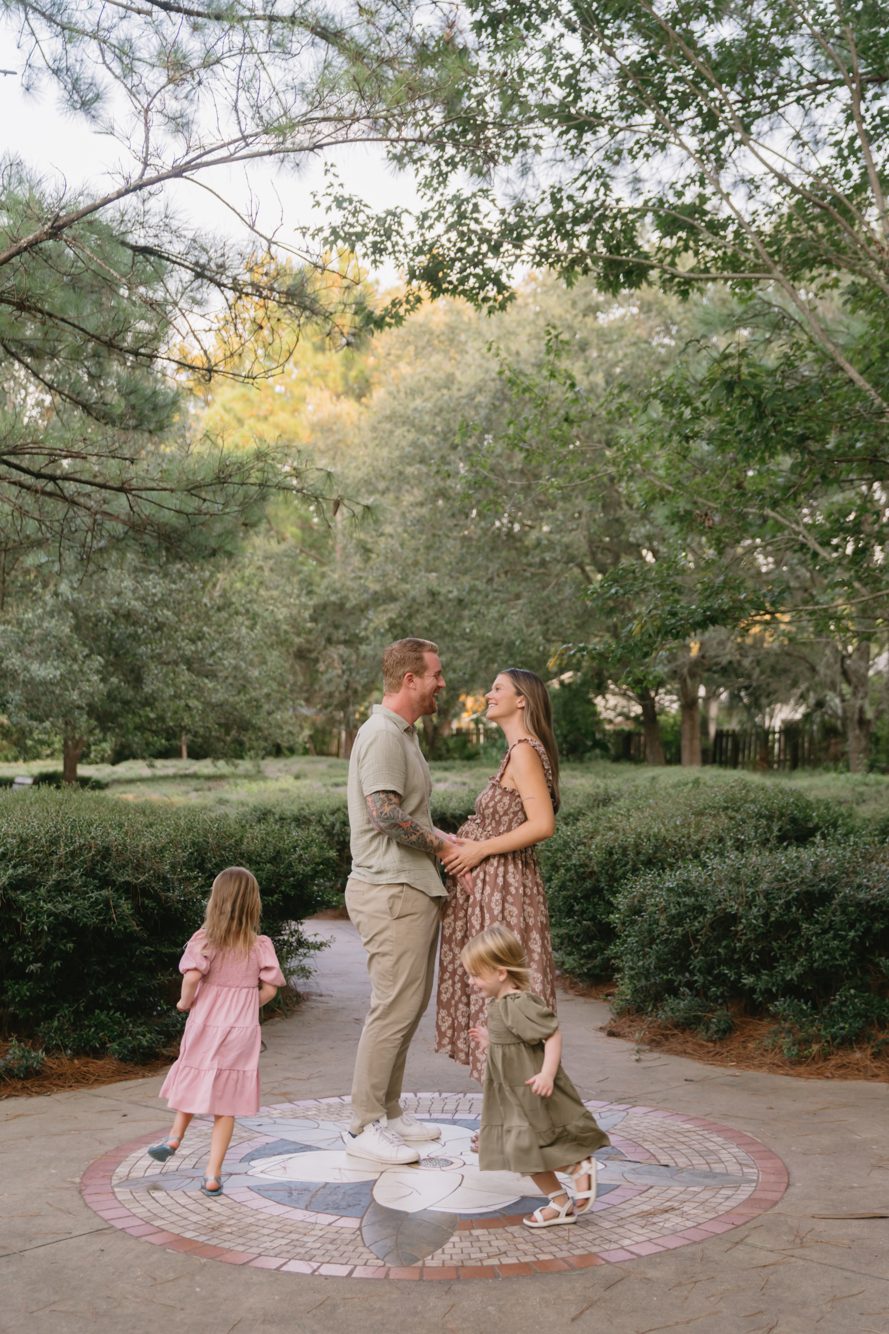 Expecting couple laughing as their daughters dance around them on the garden path at Bird Island Park by Lee Photo + Video