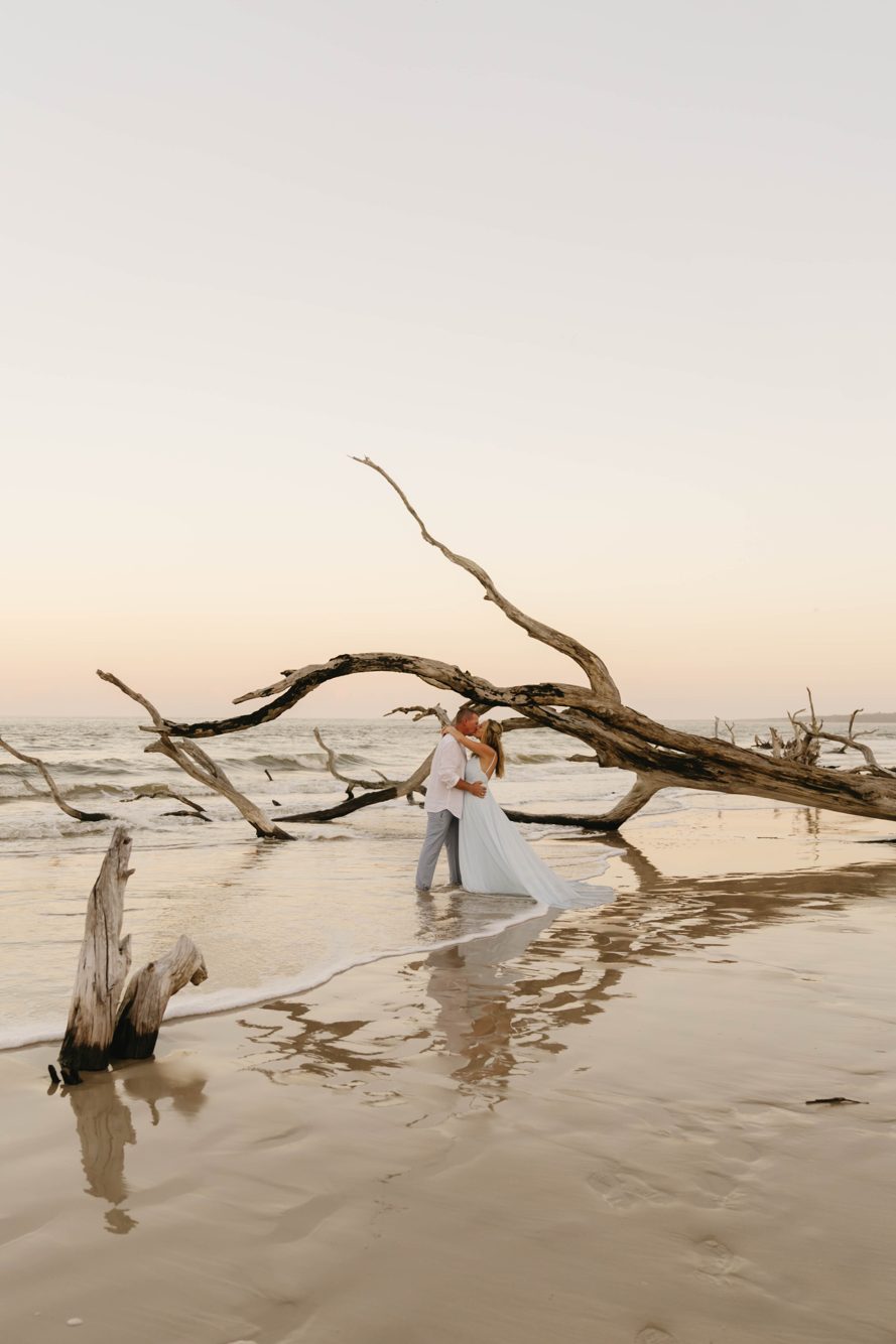 Couple kissing in the surf beneath an arching driftwood tree at sunset, Boneyard Beach on Big Talbot Island FL by Lee Photo + Video