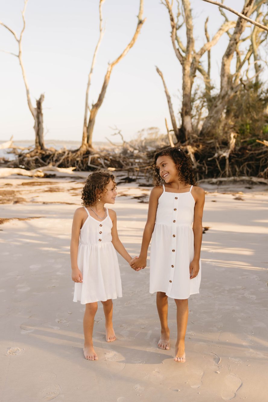 Two sisters holding hands among the sun-bleached driftwood at Boneyard Beach, Jacksonville FL by Lee Photo + Video