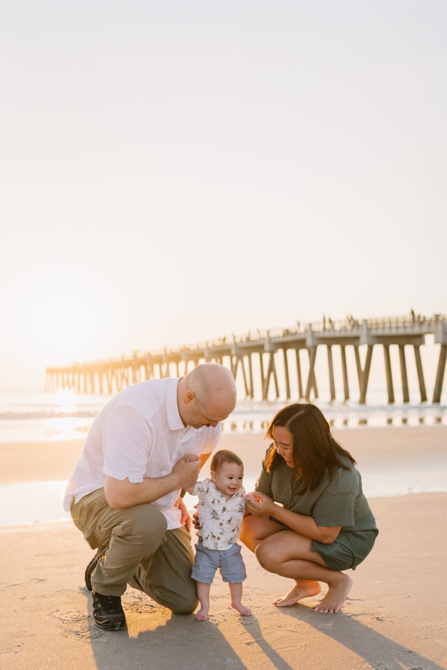 Parents helping baby take steps in the sand at sunrise with the Jacksonville Beach Pier behind them, Florida