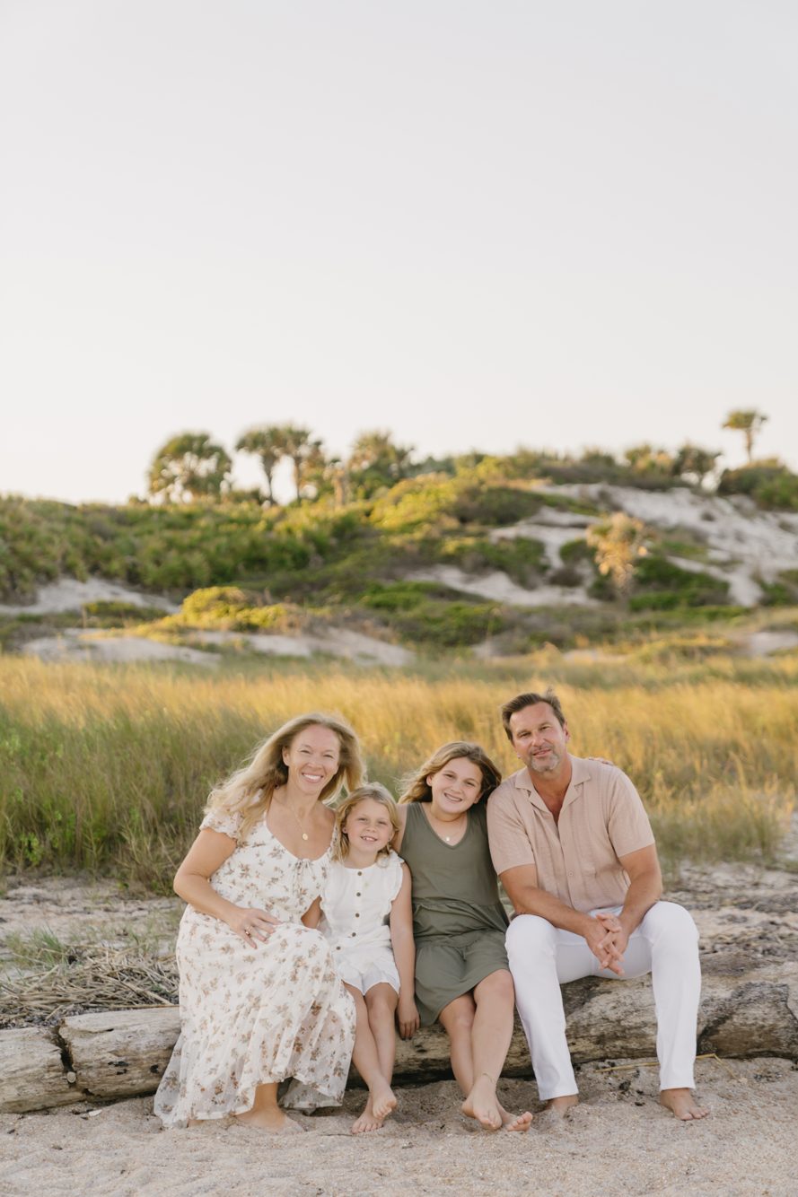 Family of four sitting on driftwood with the grassy dunes behind them at North Beach Guana State Park in Ponte Vedra Beach FL by Lee Photo + Video