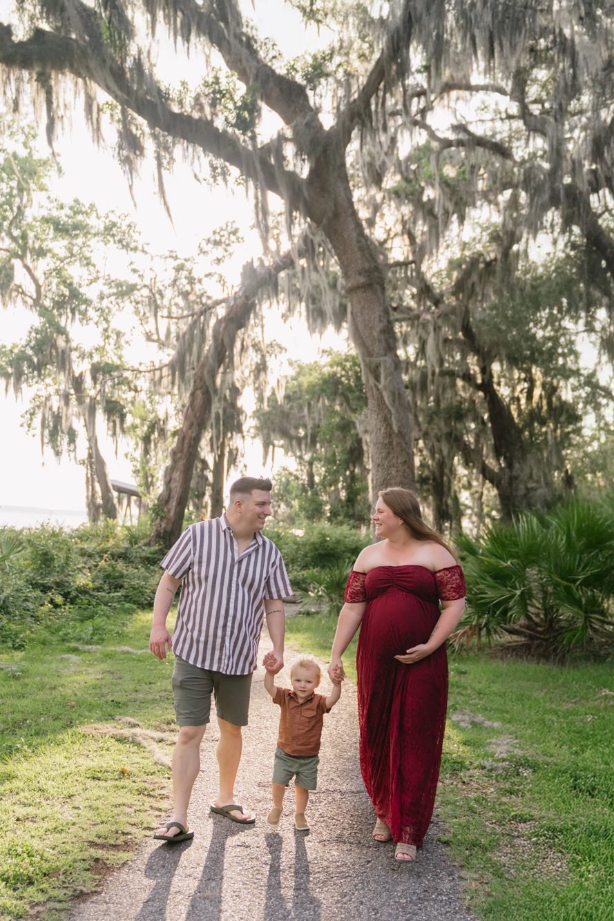 Family walking hand in hand with their toddler beneath moss-draped oaks at Riverfront Park in St. Johns County FL by Lee Photo + Video